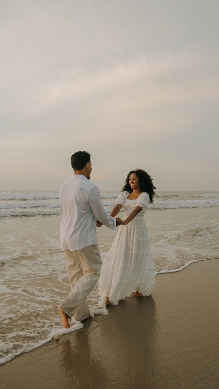 Couple in white clothing holding hands while walking on a sandy beach at sunset.