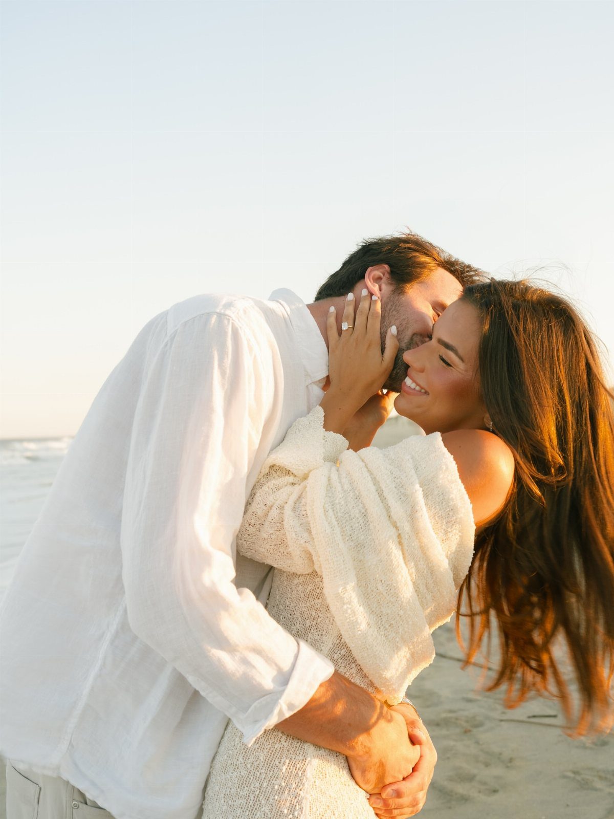 Couple in white clothing embracing and kissing on a sunny sandy beach.