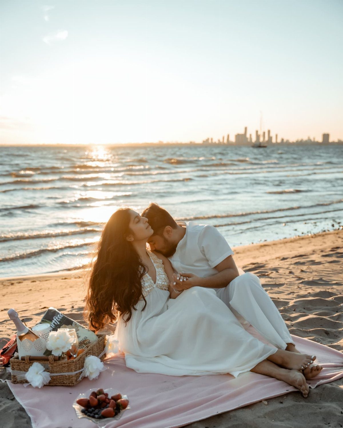 Couple sitting on a pink blanket with a picnic basket on a beach.