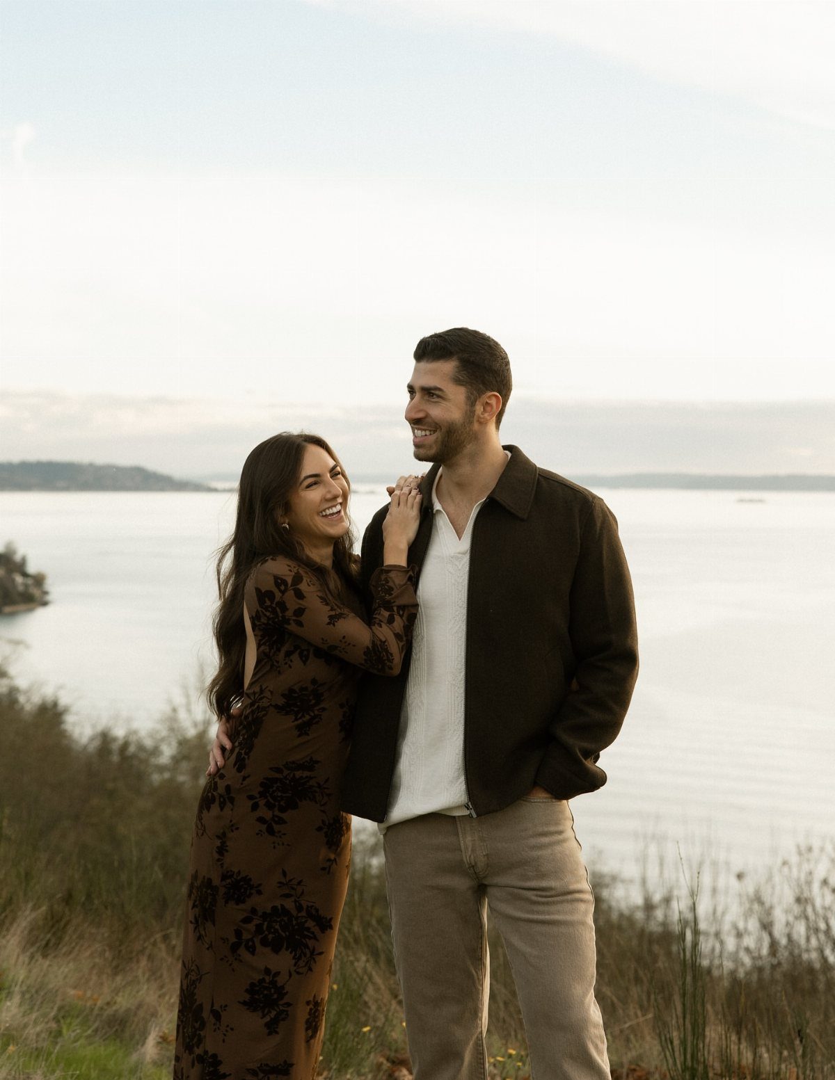 Smiling couple posing on a grassy cliff overlooking a calm ocean bay.