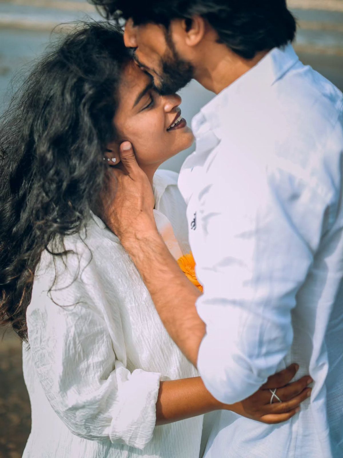 Couple in white clothing embracing with the man kissing the woman's forehead.