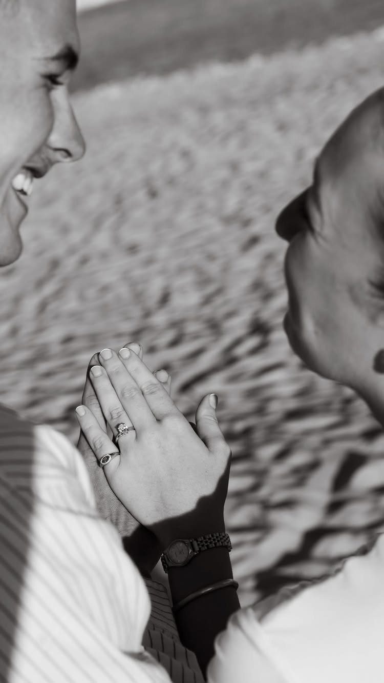 Couple facing each other with a close-up of an engagement ring on a beach