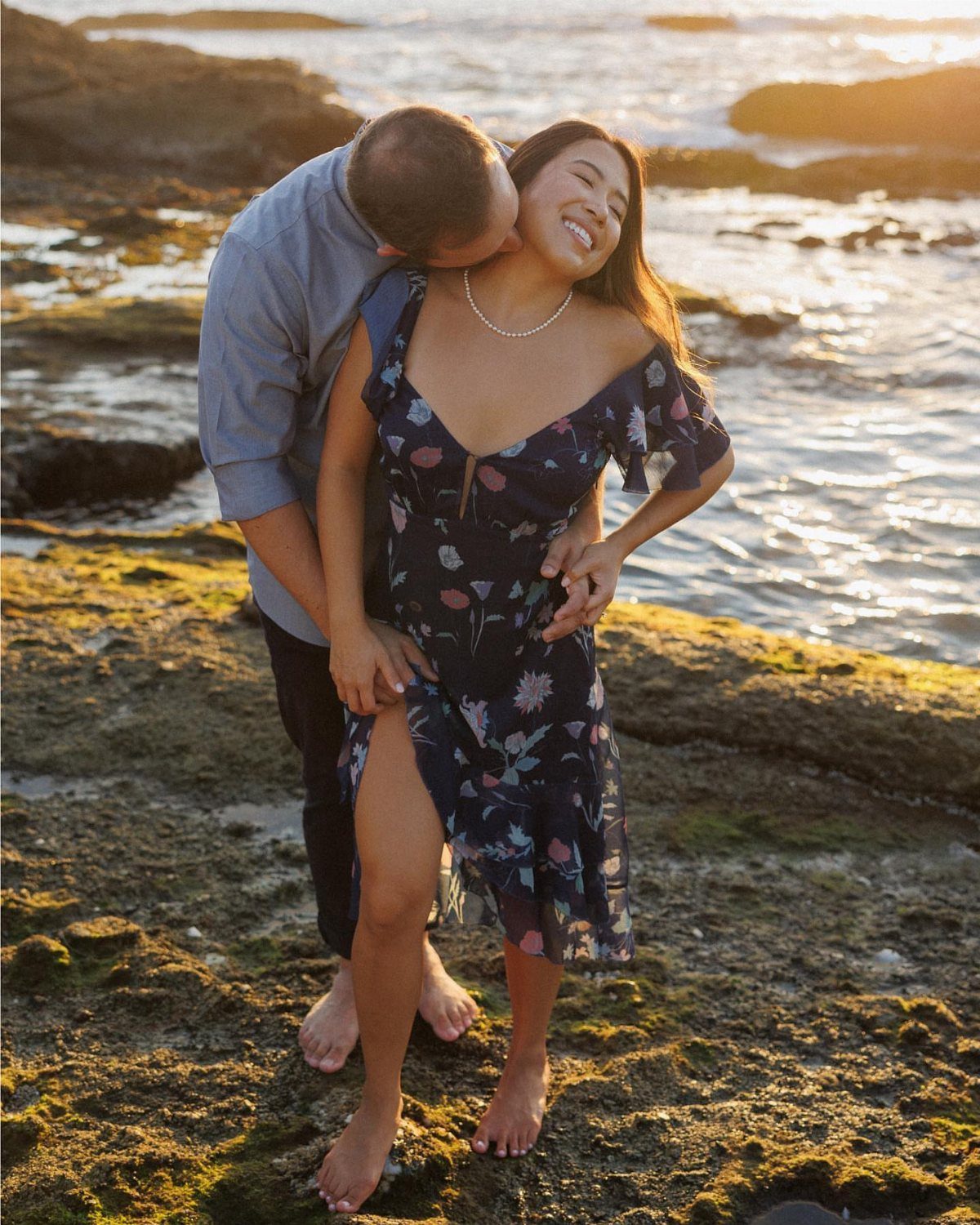 Couple embracing on a rocky beach at sunset, man kissing woman's cheek.
