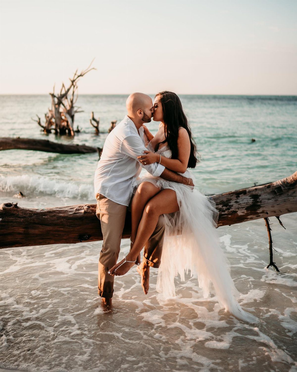 Couple kissing on a beach while sitting on a large piece of driftwood.
