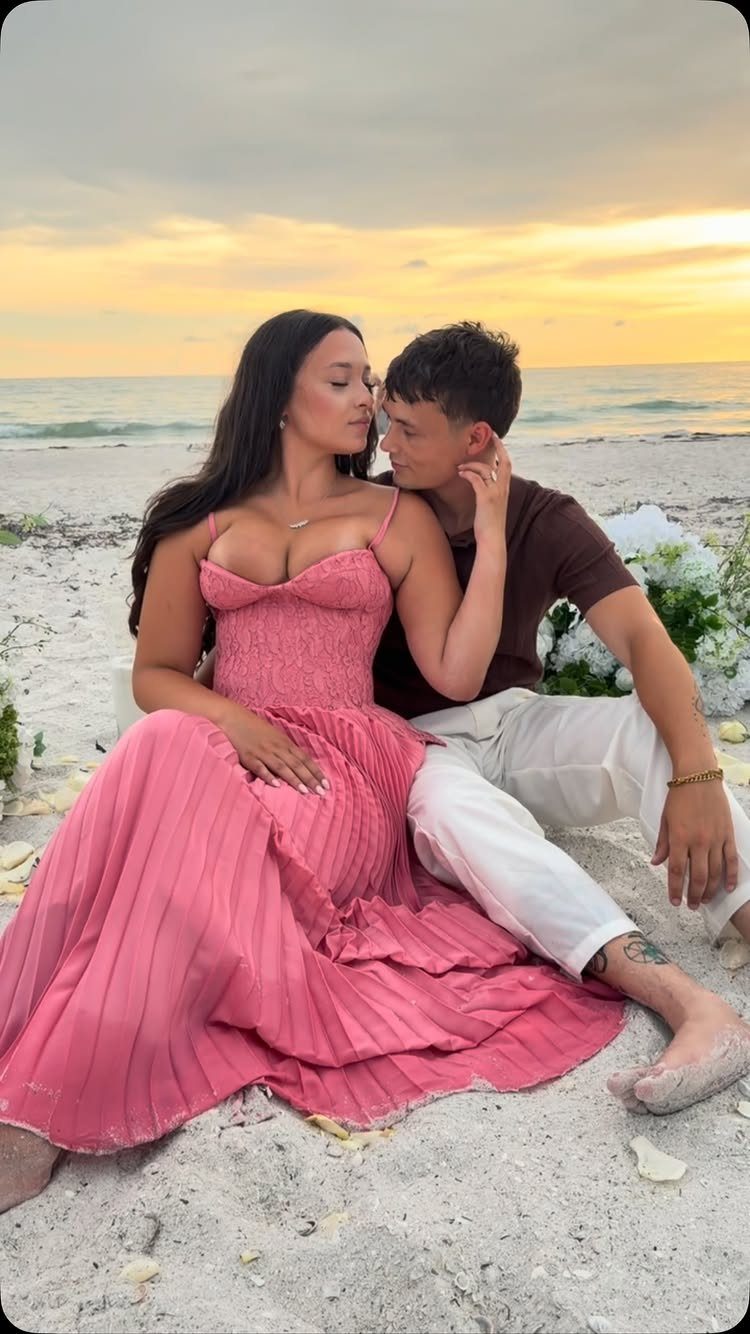 Couple in pink dress and white pants sitting on a beach at sunset.