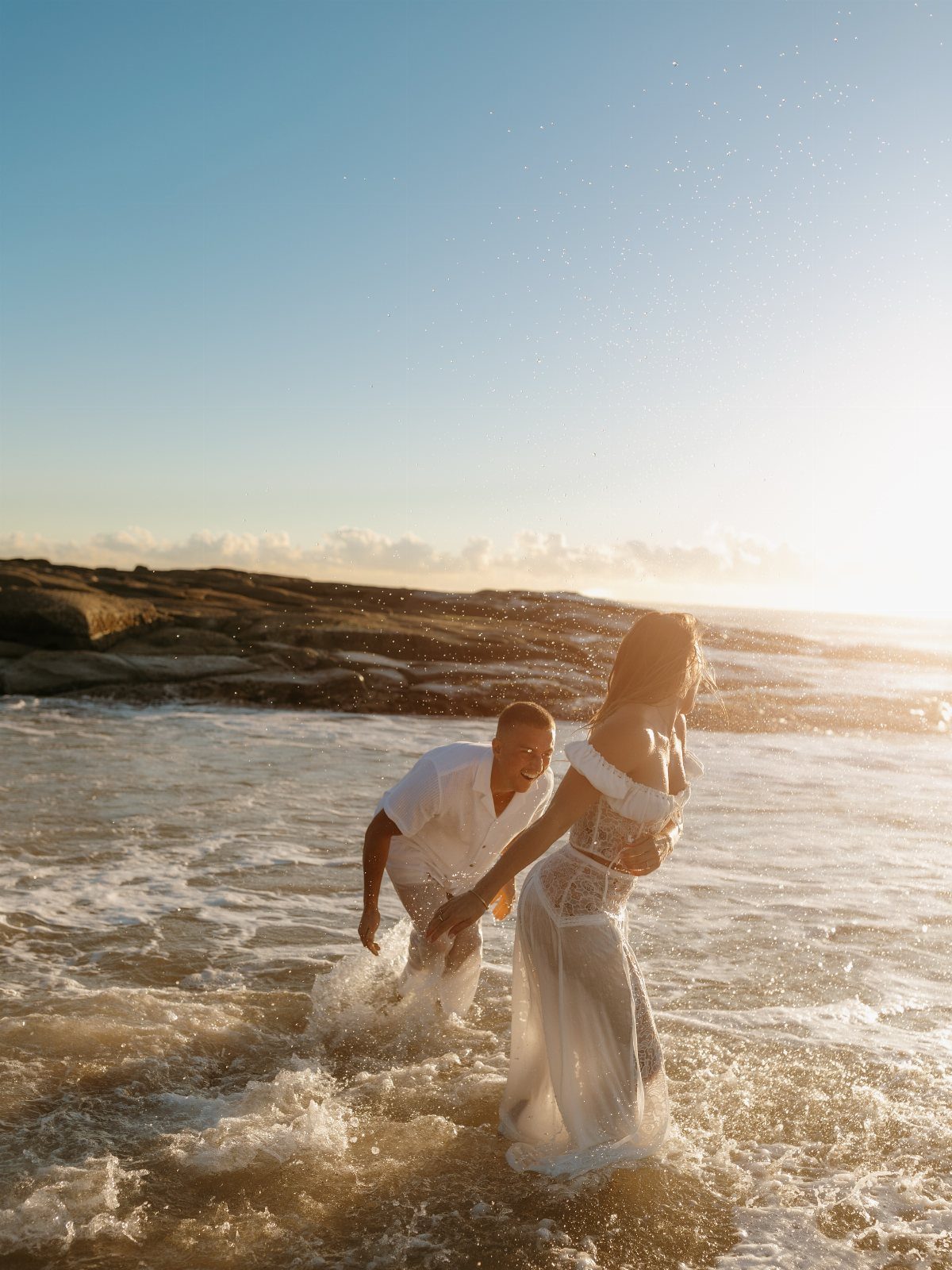 Couple in white clothing splashing in ocean waves during a beach sunset.