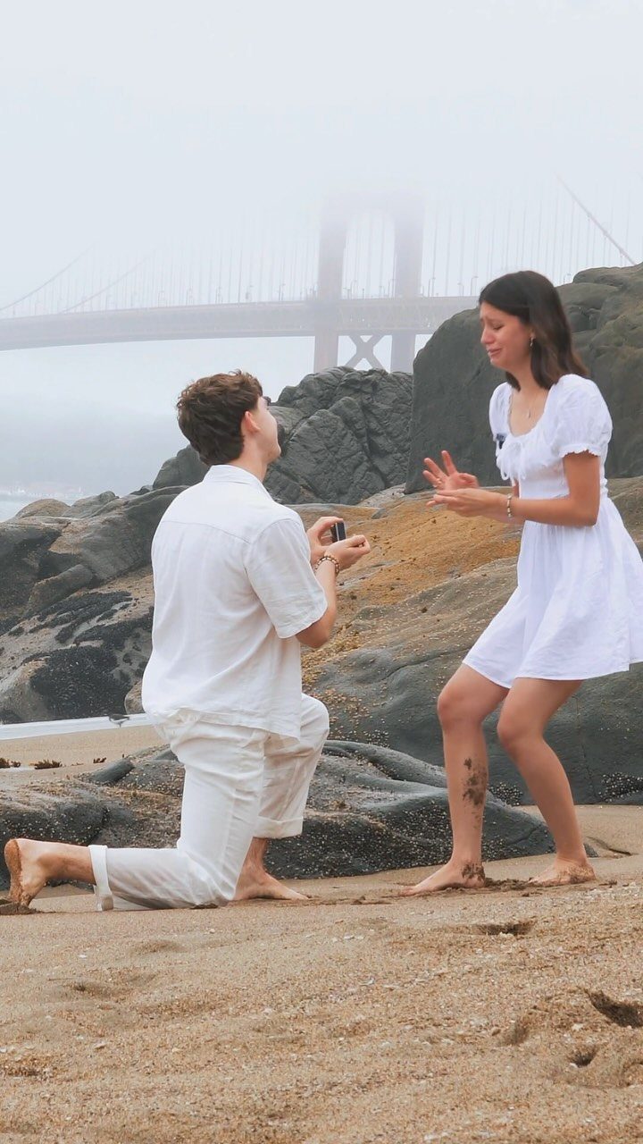 Man proposing to a woman on a sandy beach with a foggy bridge background