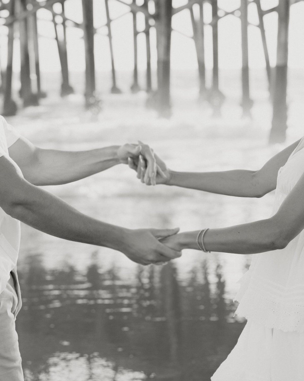 Couple holding hands on a beach with a wooden pier in the background.