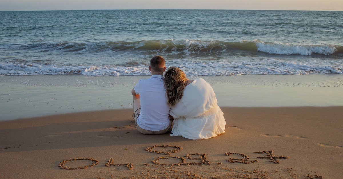 Couple sitting on beach with a date and heart written in the sand.