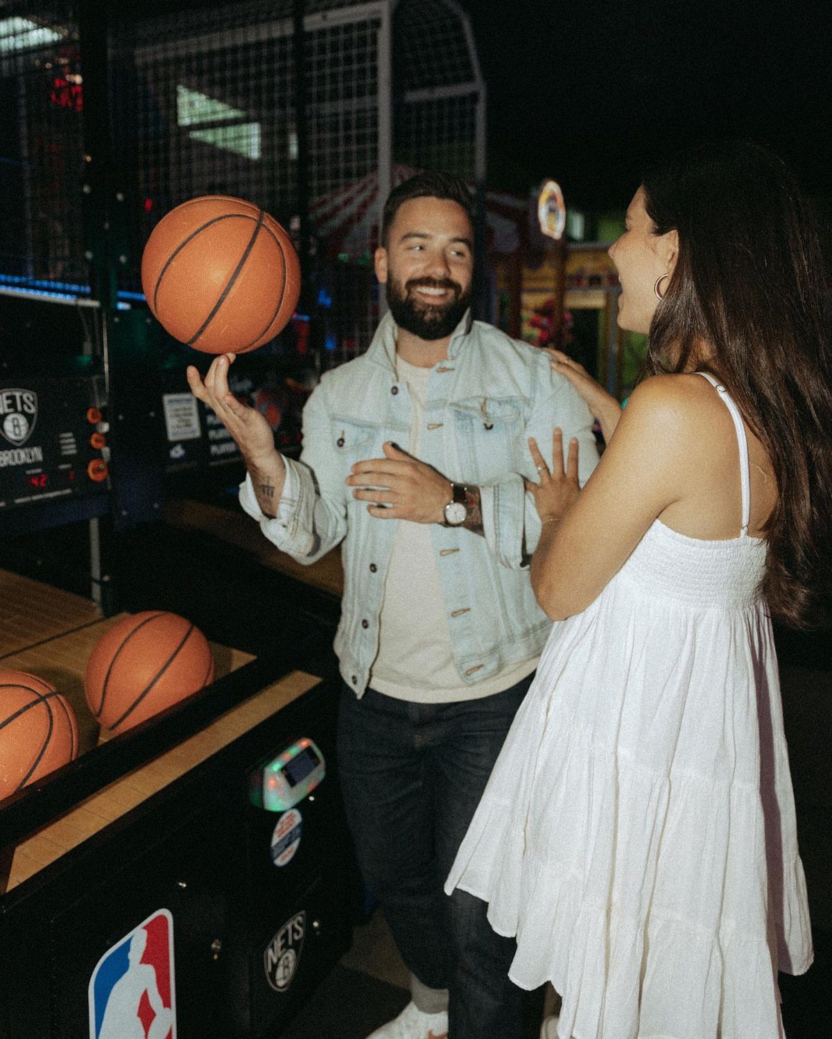 Couple posing by a basketball arcade game in a dimly lit barcade venue.