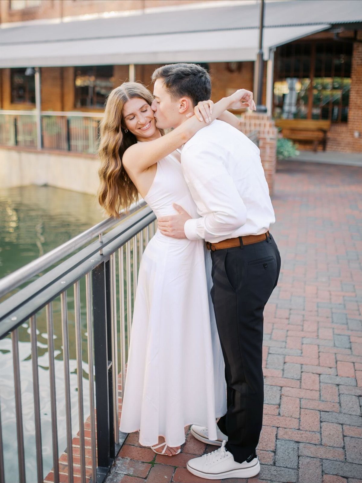Couple embracing on a red brick walkway next to water and brick buildings.