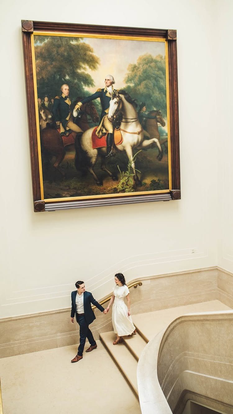 Couple holding hands walking down a gallery staircase beneath a large framed painting.