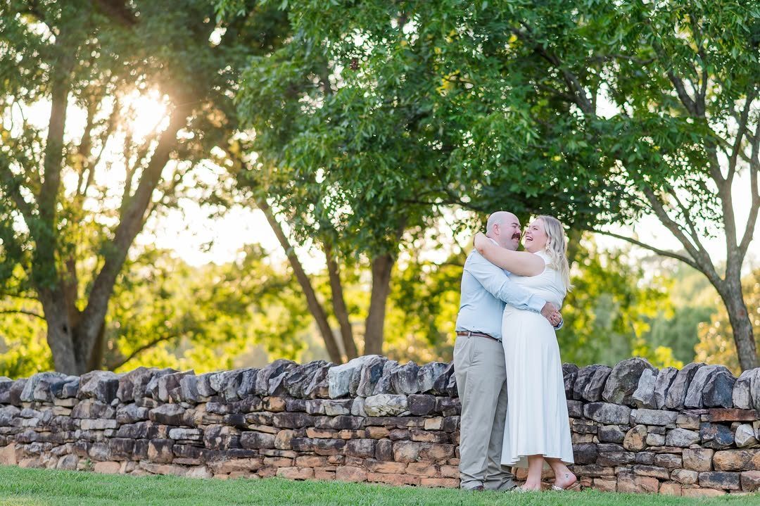 Smiling couple embracing beside a rustic stone wall under large green oak trees.