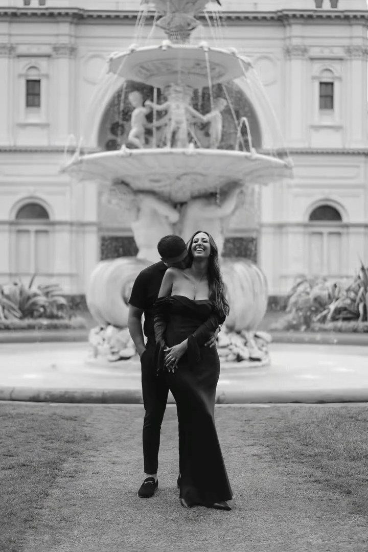 Black couple in formal attire embracing before a large ornate stone fountain.