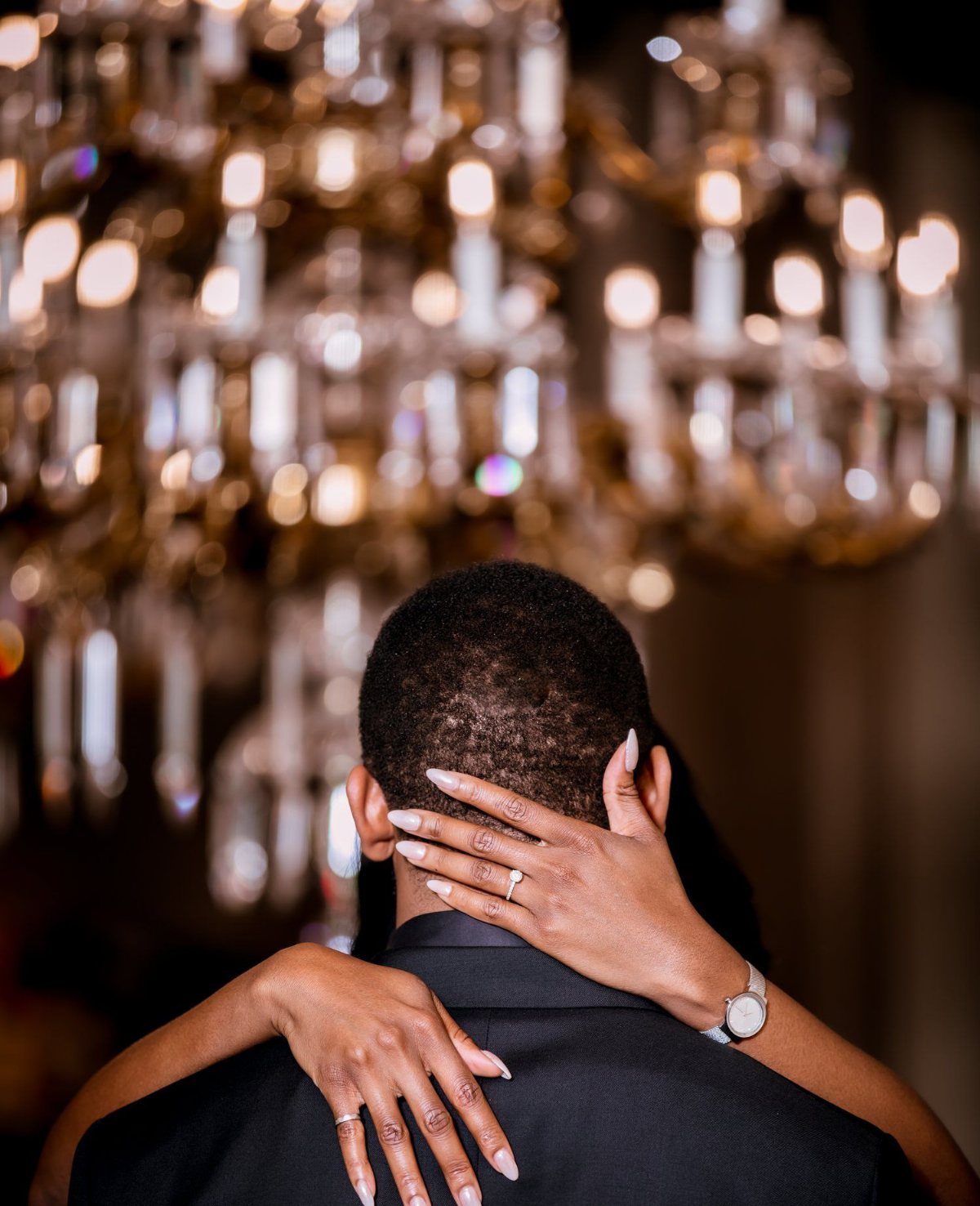 Black couple embracing with a focus on the engagement ring and gold chandelier.
