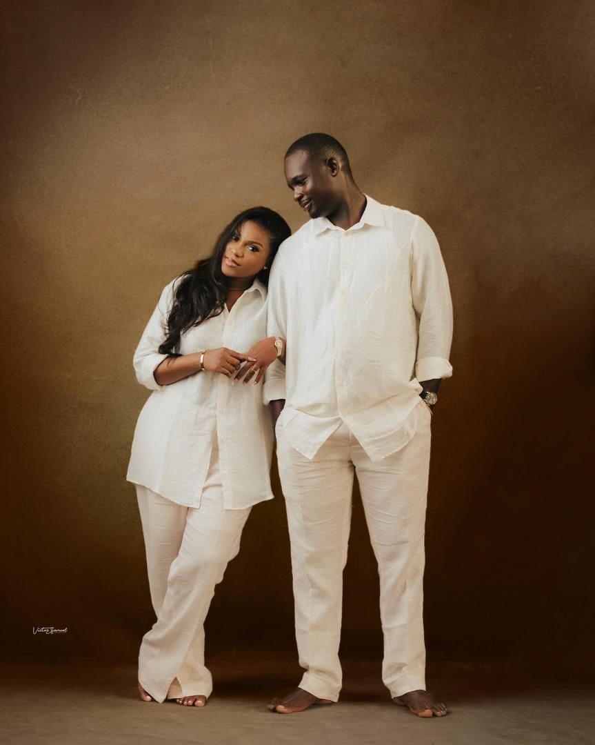 Black couple in matching white linen outfits posing against a brown studio background