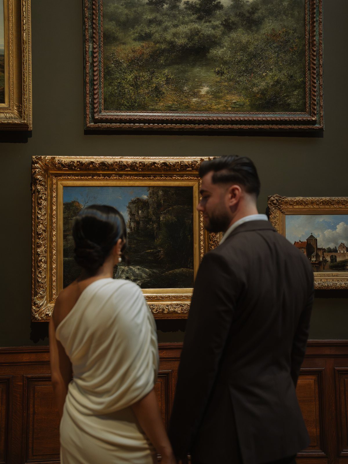 Couple in formal attire viewing landscape paintings inside an art museum gallery
