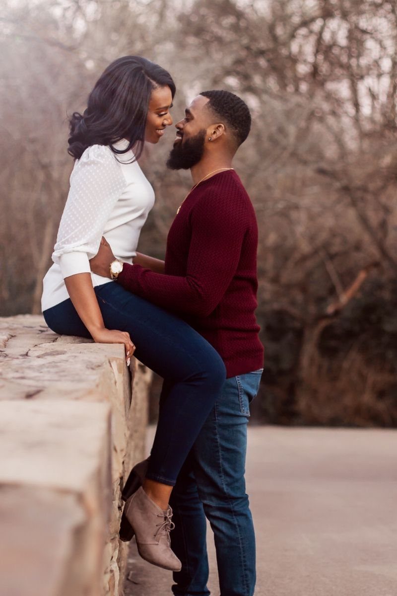 Black couple smiling and embracing outdoors by a stone wall with bare trees.