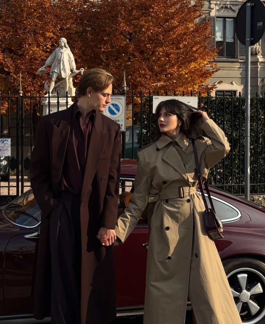 Couple in burgundy and tan coats holding hands by a maroon car in autumn.