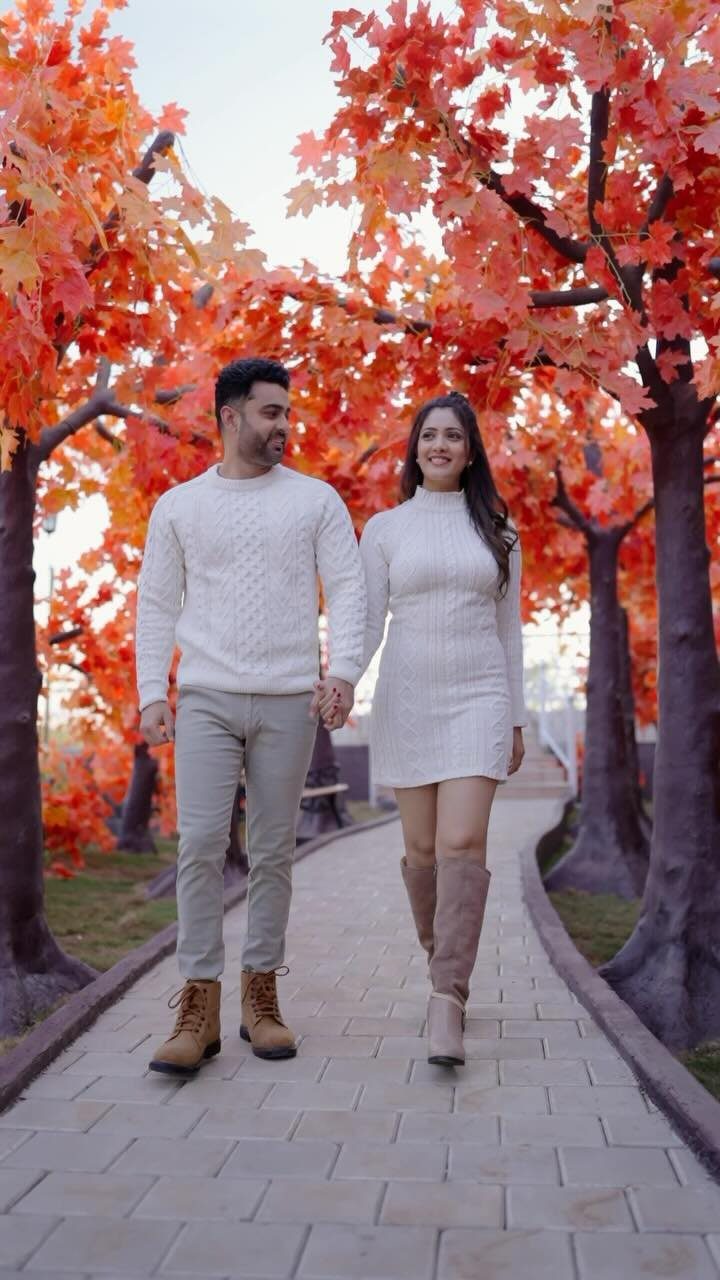 Couple in white knit outfits walking through a path of orange autumn trees.