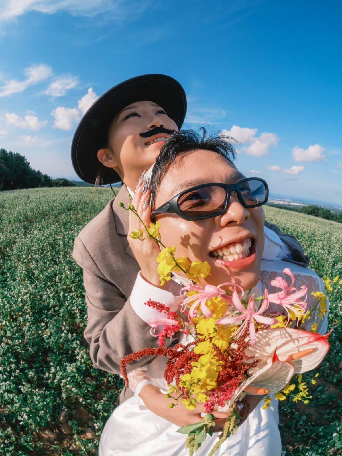 Couple posing in a field, one wearing a fake mustache and the other sunglasses.