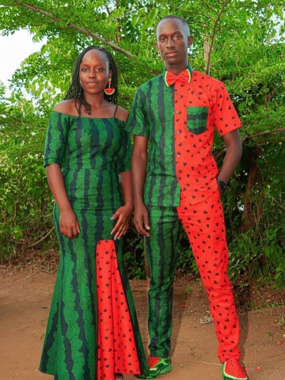 Couple wearing matching watermelon-themed clothing standing outdoors against a backdrop of green trees.