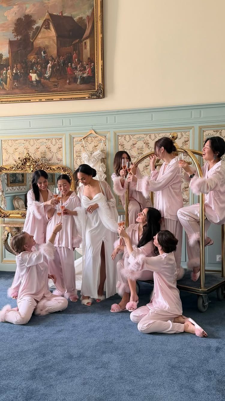 Bride and bridesmaids in robes laughing and toasting with champagne glasses in a room.
