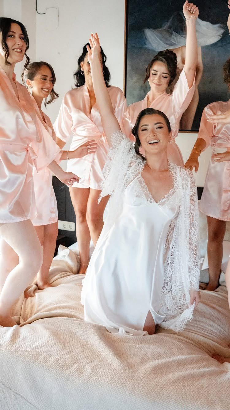 Bride in white robe cheering on a bed with bridesmaids in pink robes.