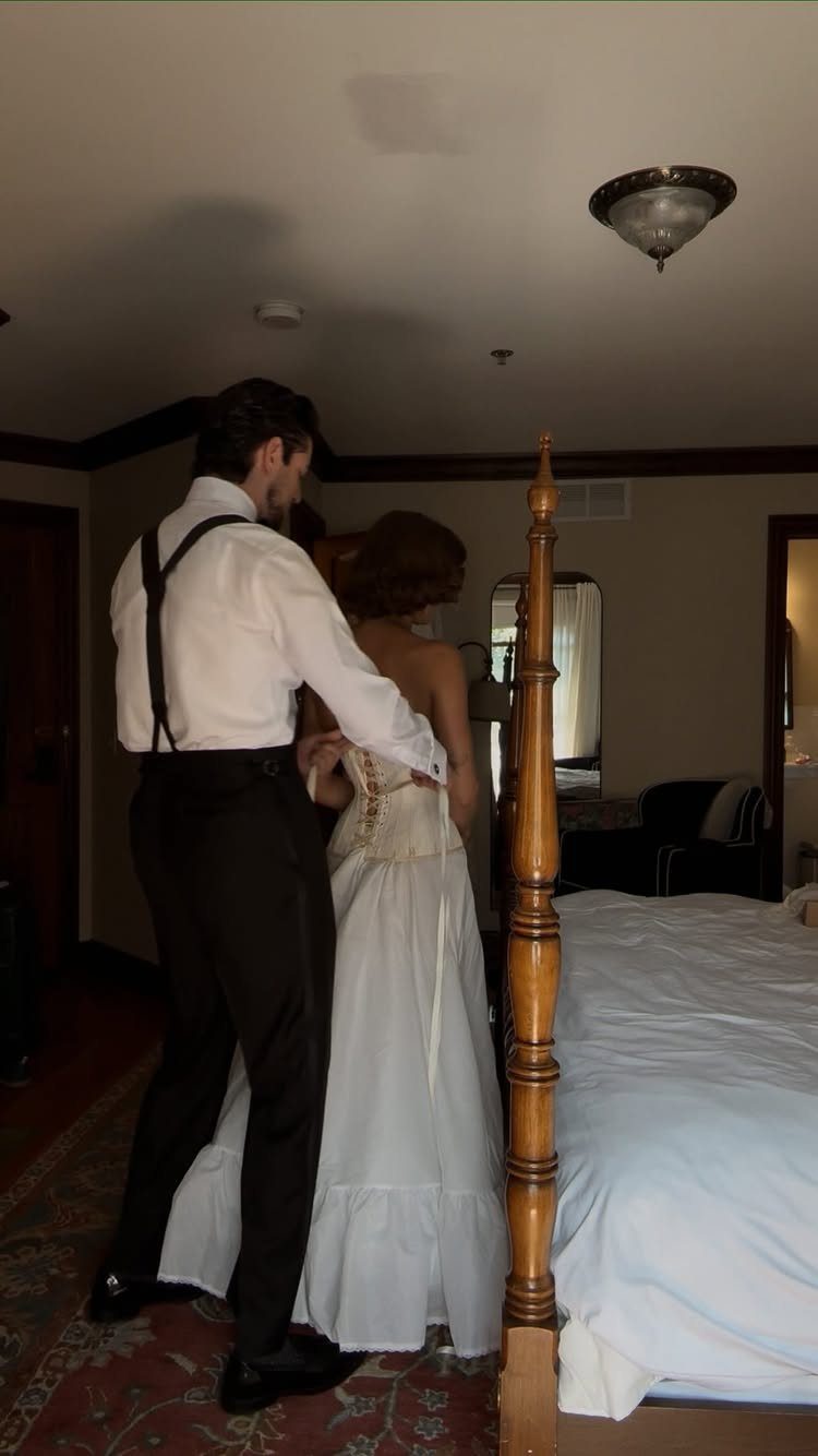Groom in suspenders helping bride with her white wedding dress in a bedroom