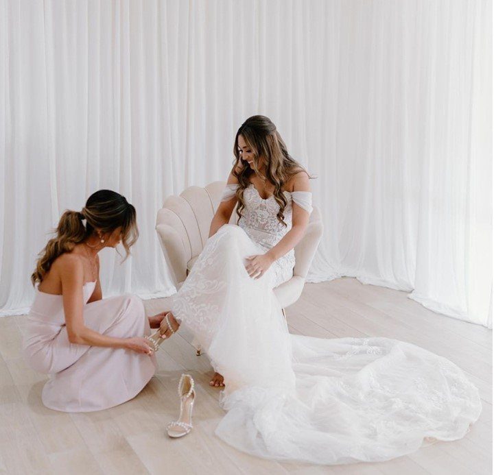 Bride in white gown getting help with her shoes from a bridesmaid.