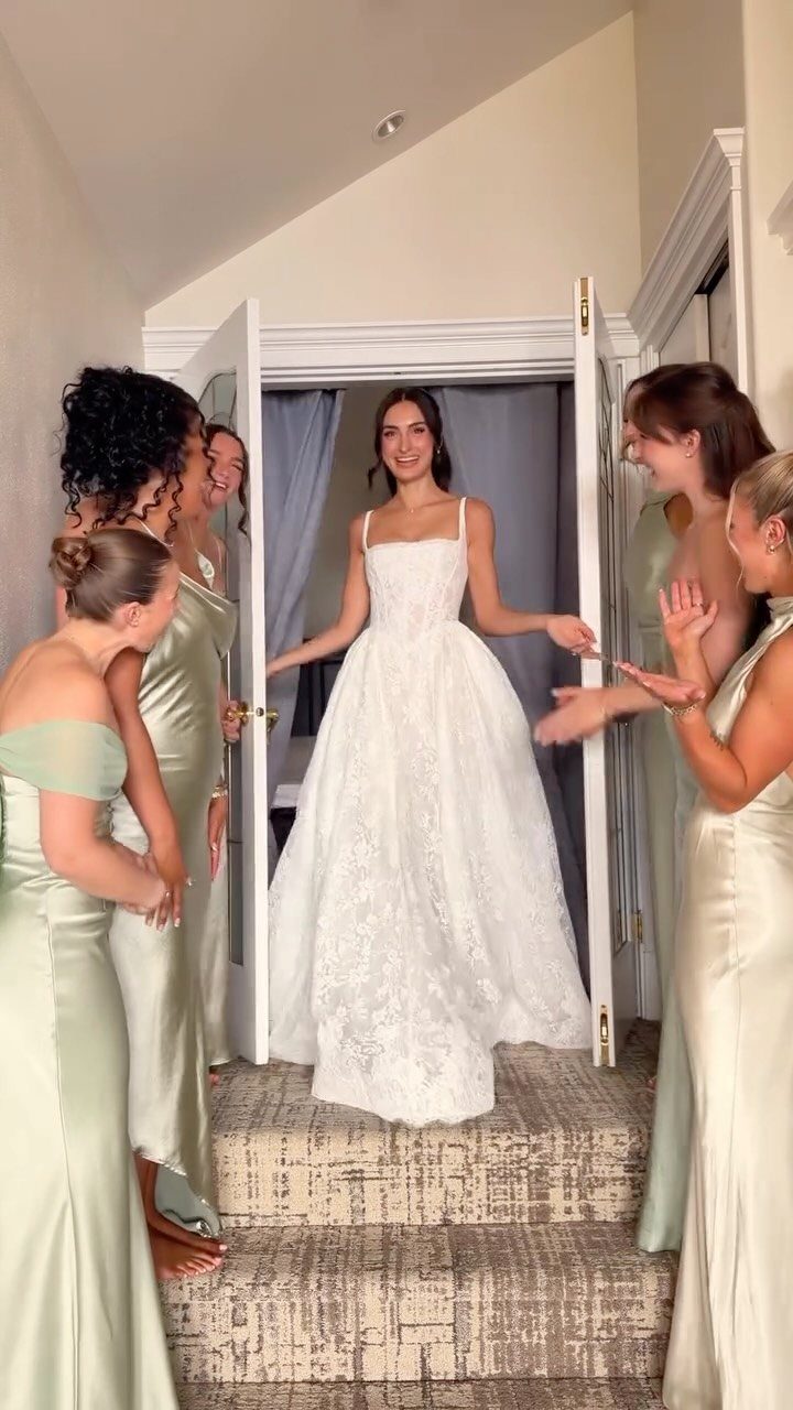 Bride in white lace gown smiling at bridesmaids in pale green satin dresses.