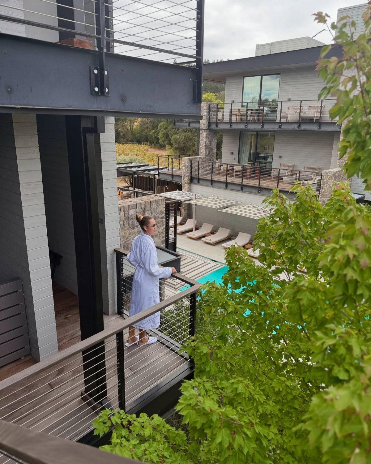 Woman in white robe on a modern balcony overlooking a pool and patio.