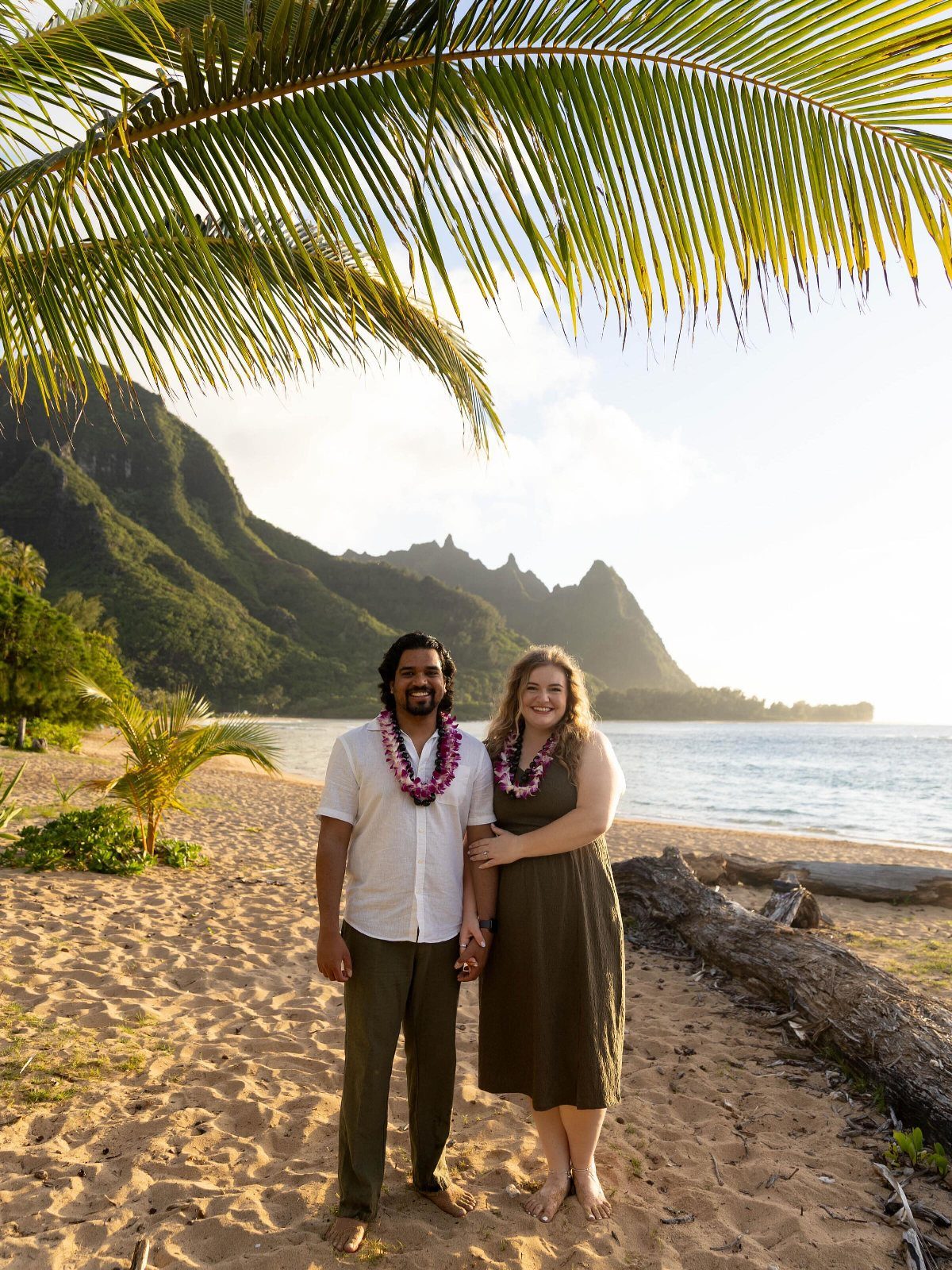 Couple wearing flower leis standing on a tropical beach with mountains in background
