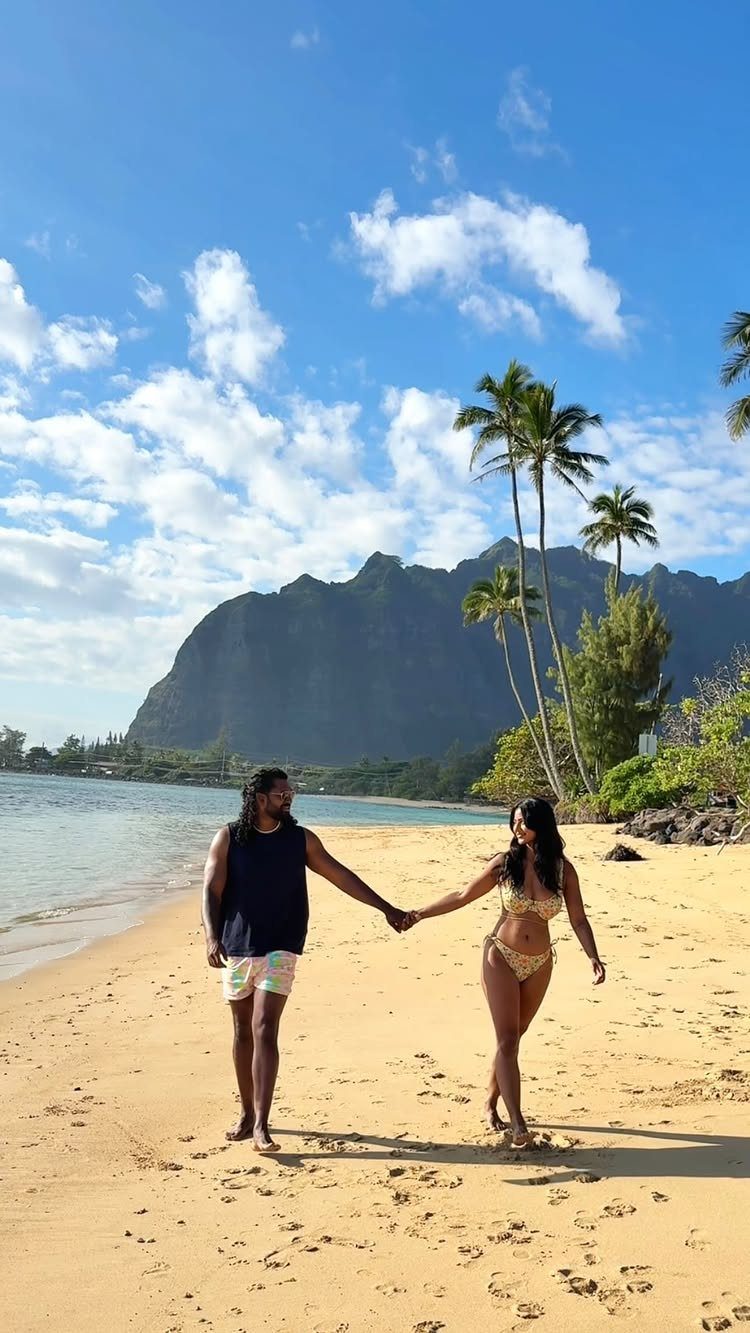 Couple holding hands walking on a tropical beach with mountains and palm trees.