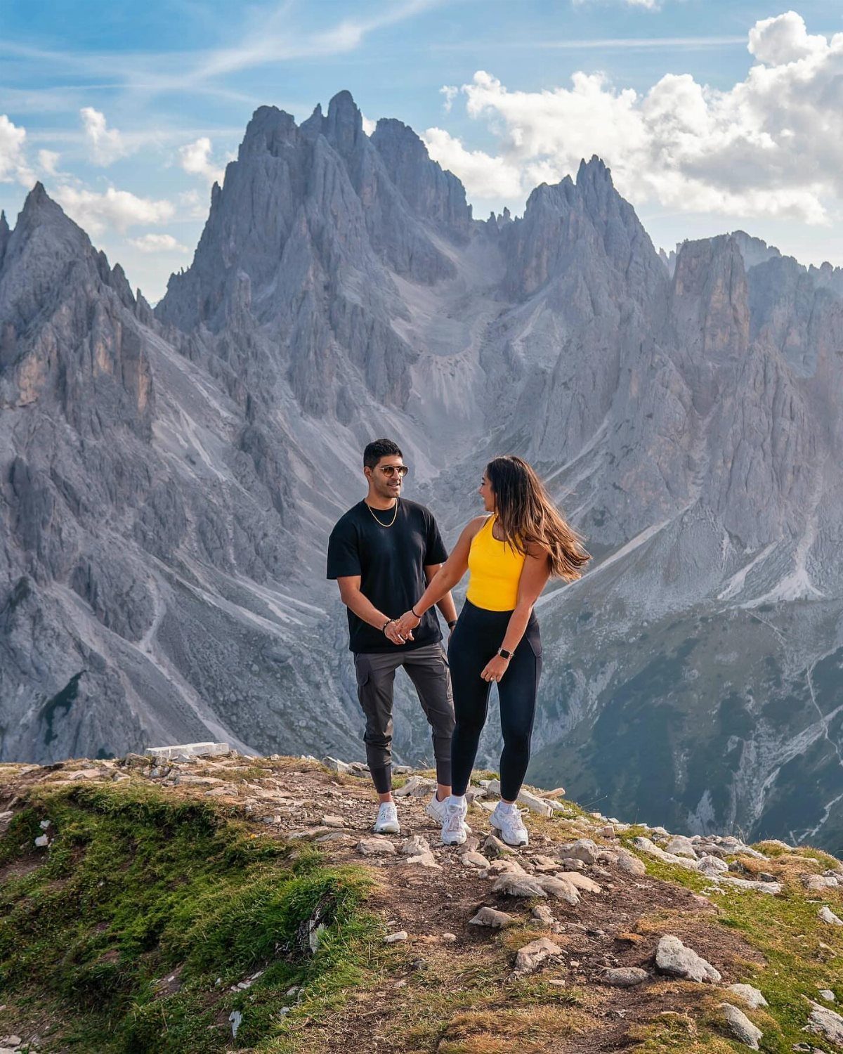 Couple holding hands on a grassy peak with jagged grey mountains behind them.