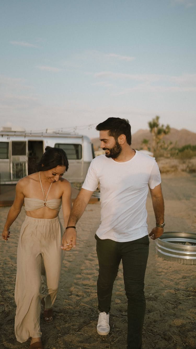 Couple holding hands and walking through a desert landscape with a vintage trailer.