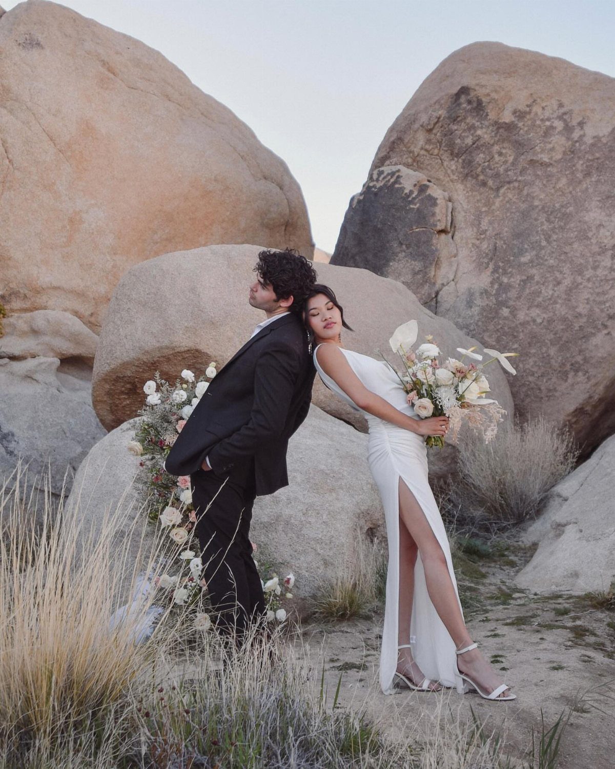 Couple in formal wedding attire posing back-to-back among large desert boulders and brush.