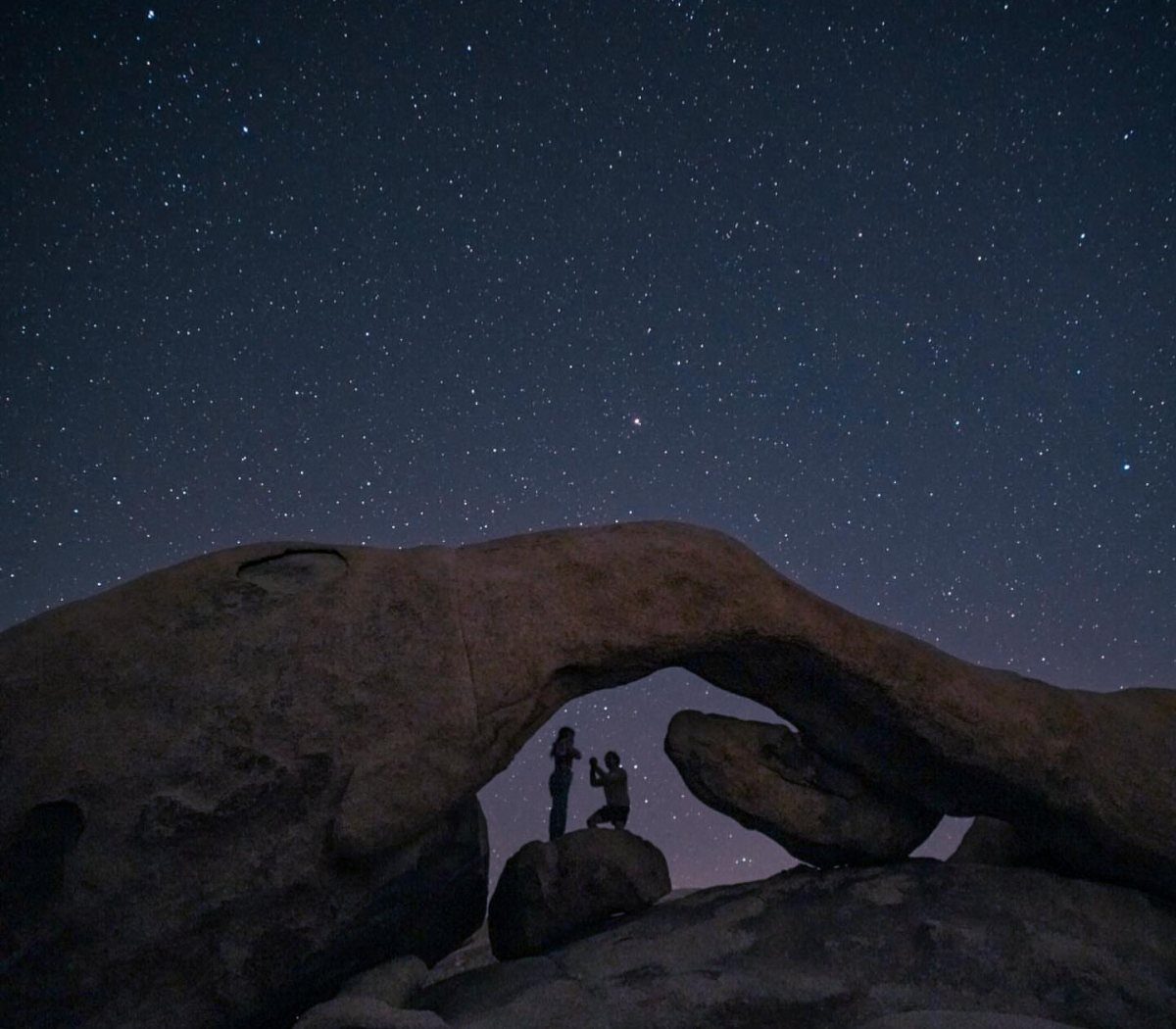 Couple silhouetted under a stone arch beneath a starry night sky in the desert