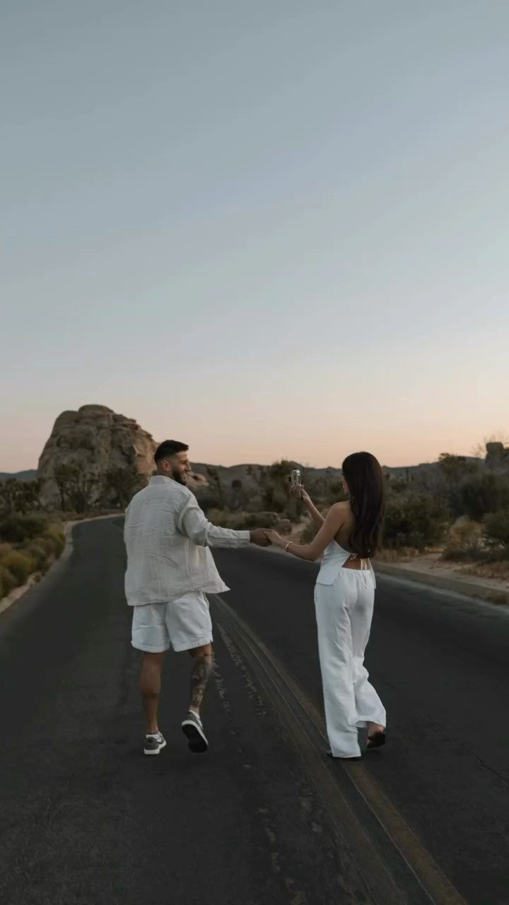 Couple in white outfits holding hands on a desert road with rocky hills.