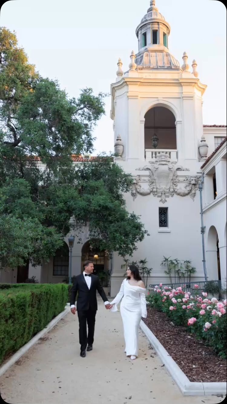 Wedding couple walking hand-in-hand in front of a white ornate tower and flowers.