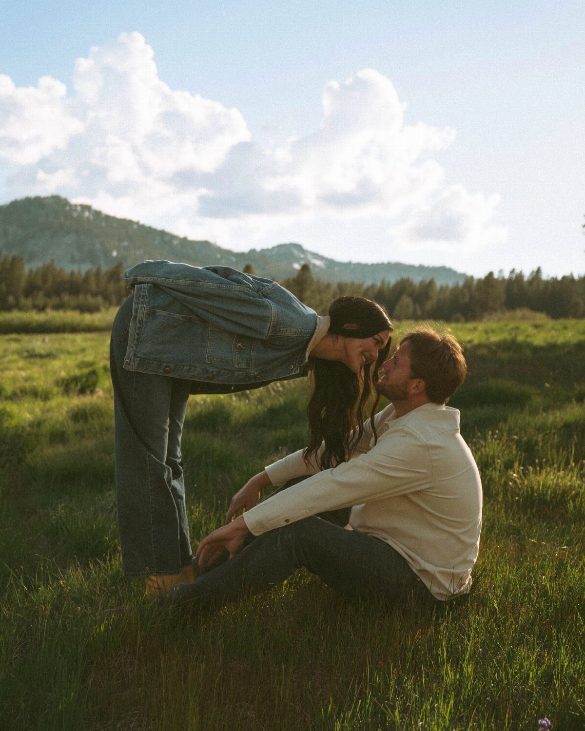 Couple interacting naturally in a grassy field with mountains under a cloudy sky.