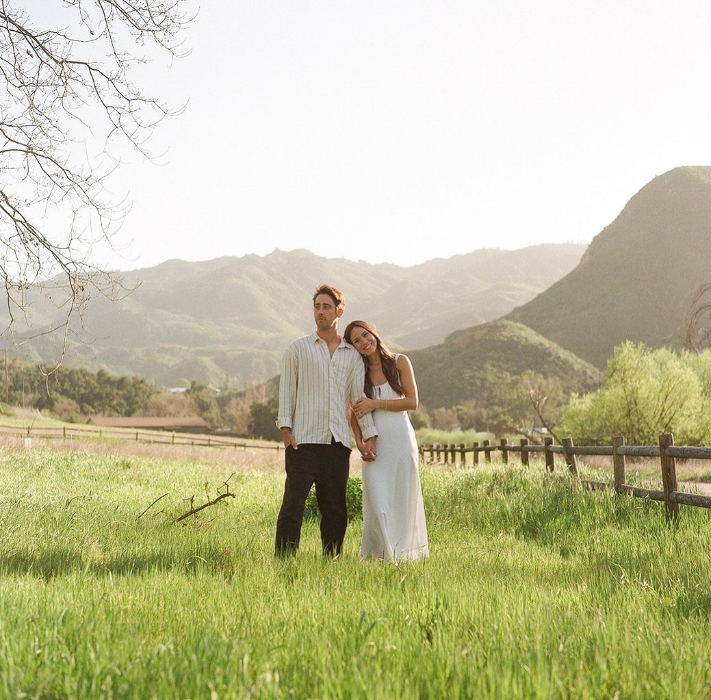 Couple holding hands in a green field with mountains under warm golden light.