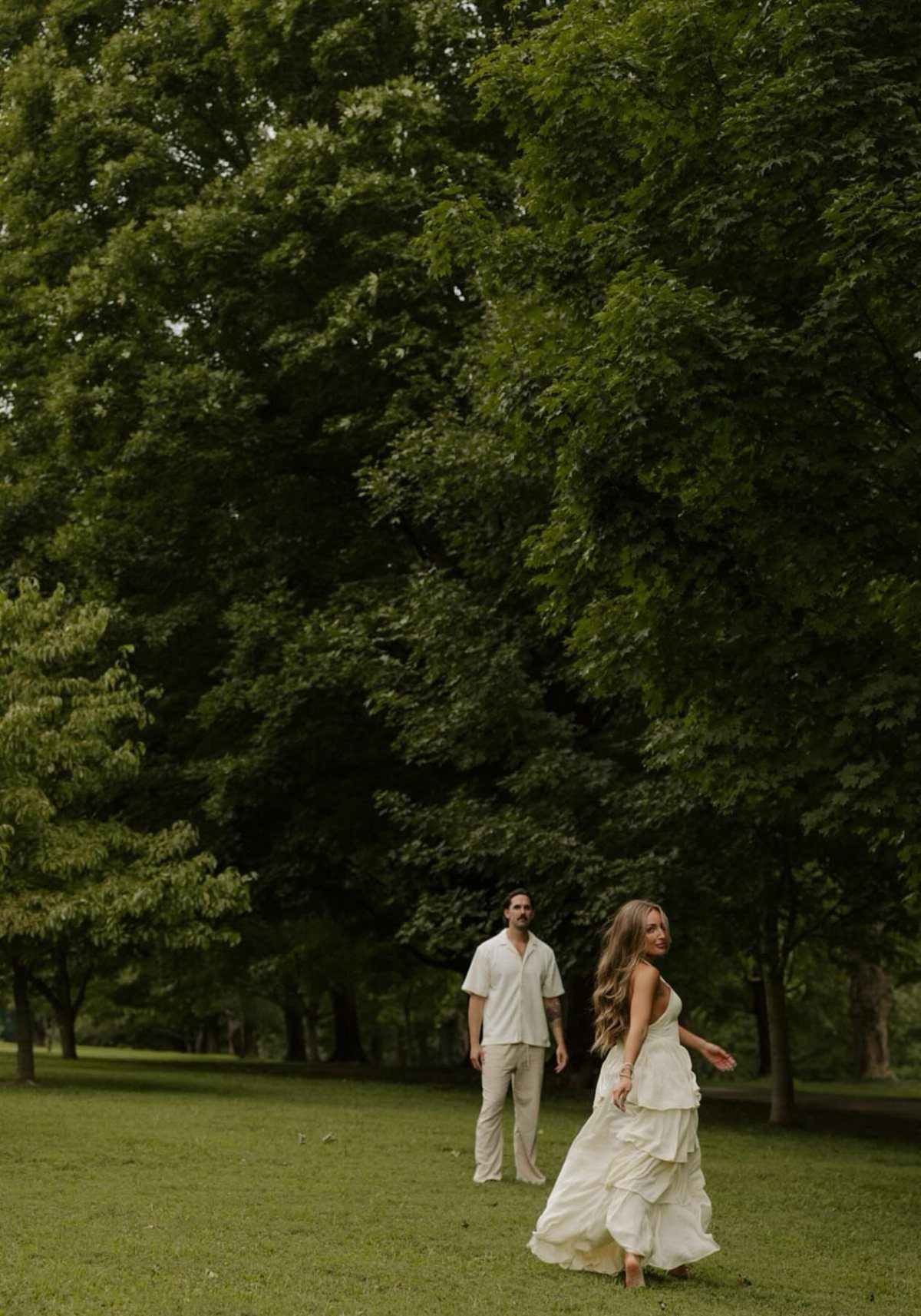 Couple in white attire standing in a grassy field with lush green trees.