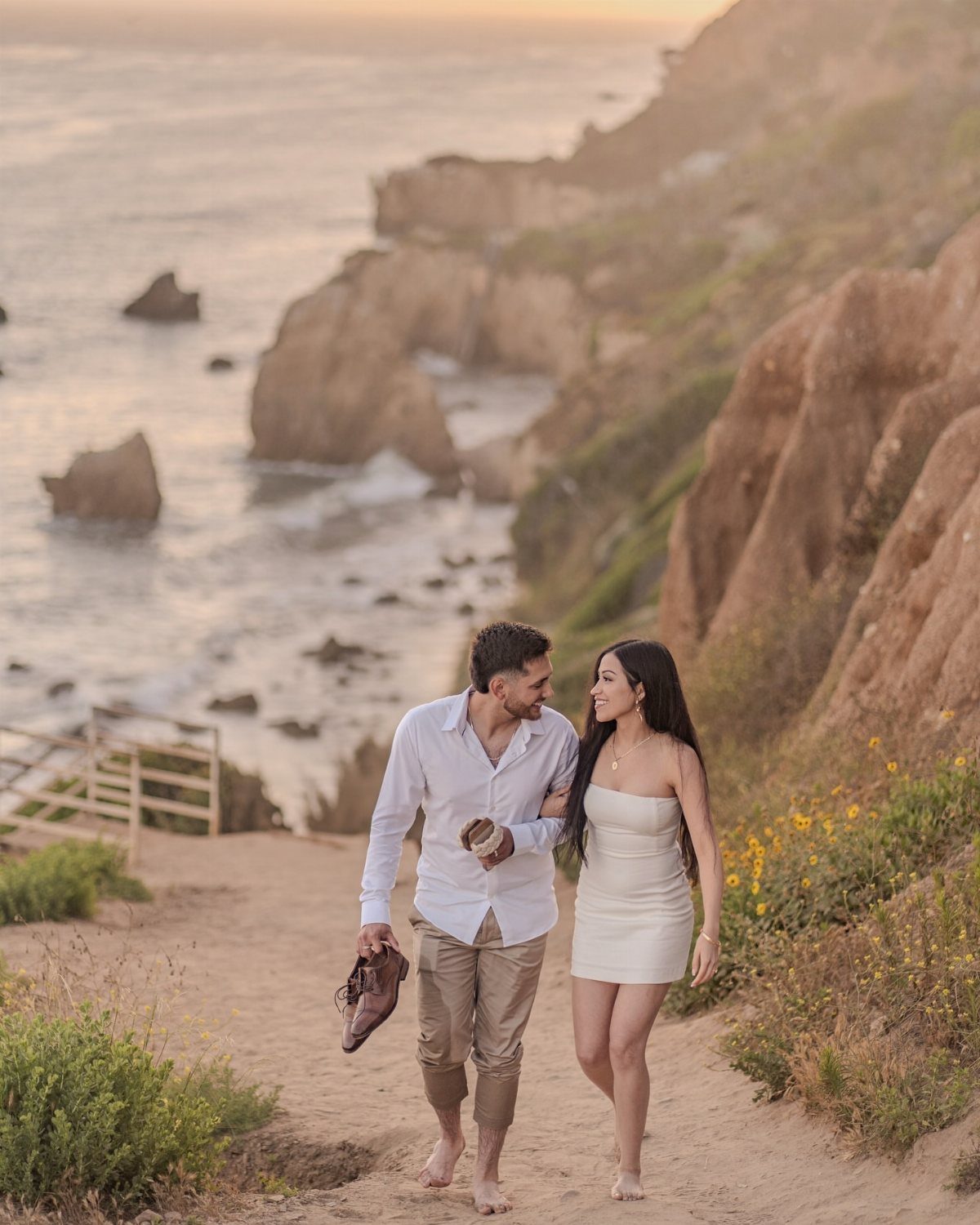 Couple in white and tan clothing walking on a coastal cliff at sunset