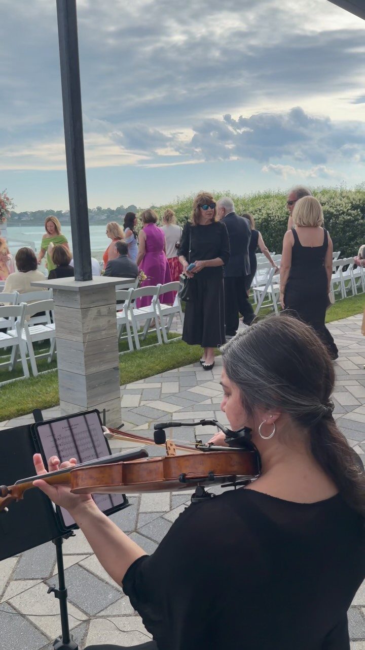 Violinist playing music for guests arriving at an outdoor wedding ceremony by the water