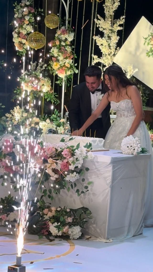 Bride and groom cutting a white wedding cake with sparks and flowers around them.
