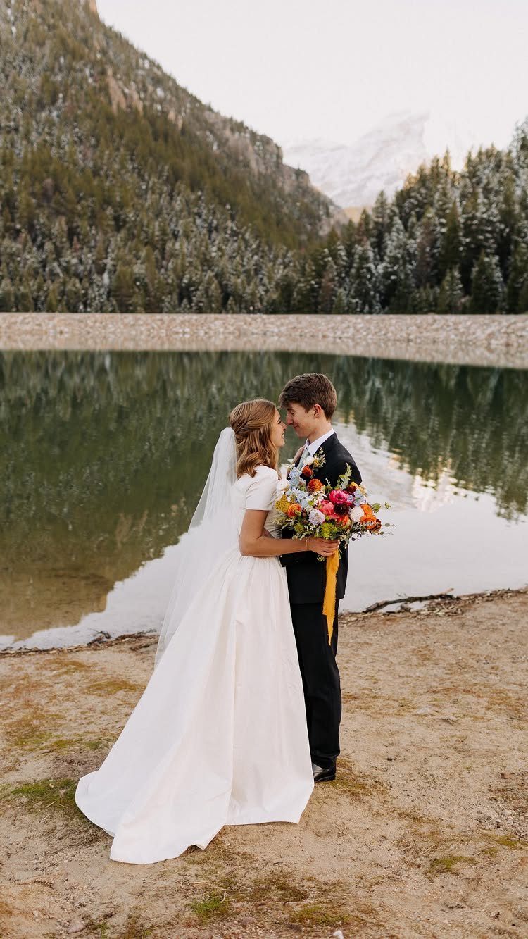 Bride and groom facing each other by a lake with mountains in background
