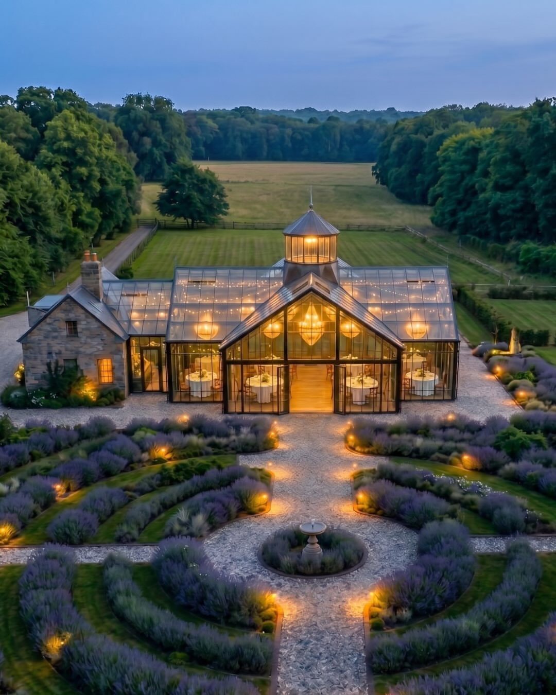 Glass conservatory building with warm lights surrounded by purple lavender gardens at dusk