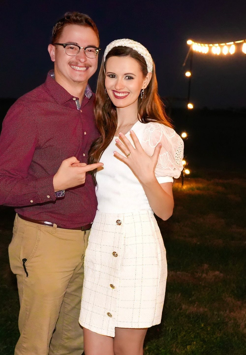 Smiling couple posing outdoors at night, woman showing off her engagement ring.