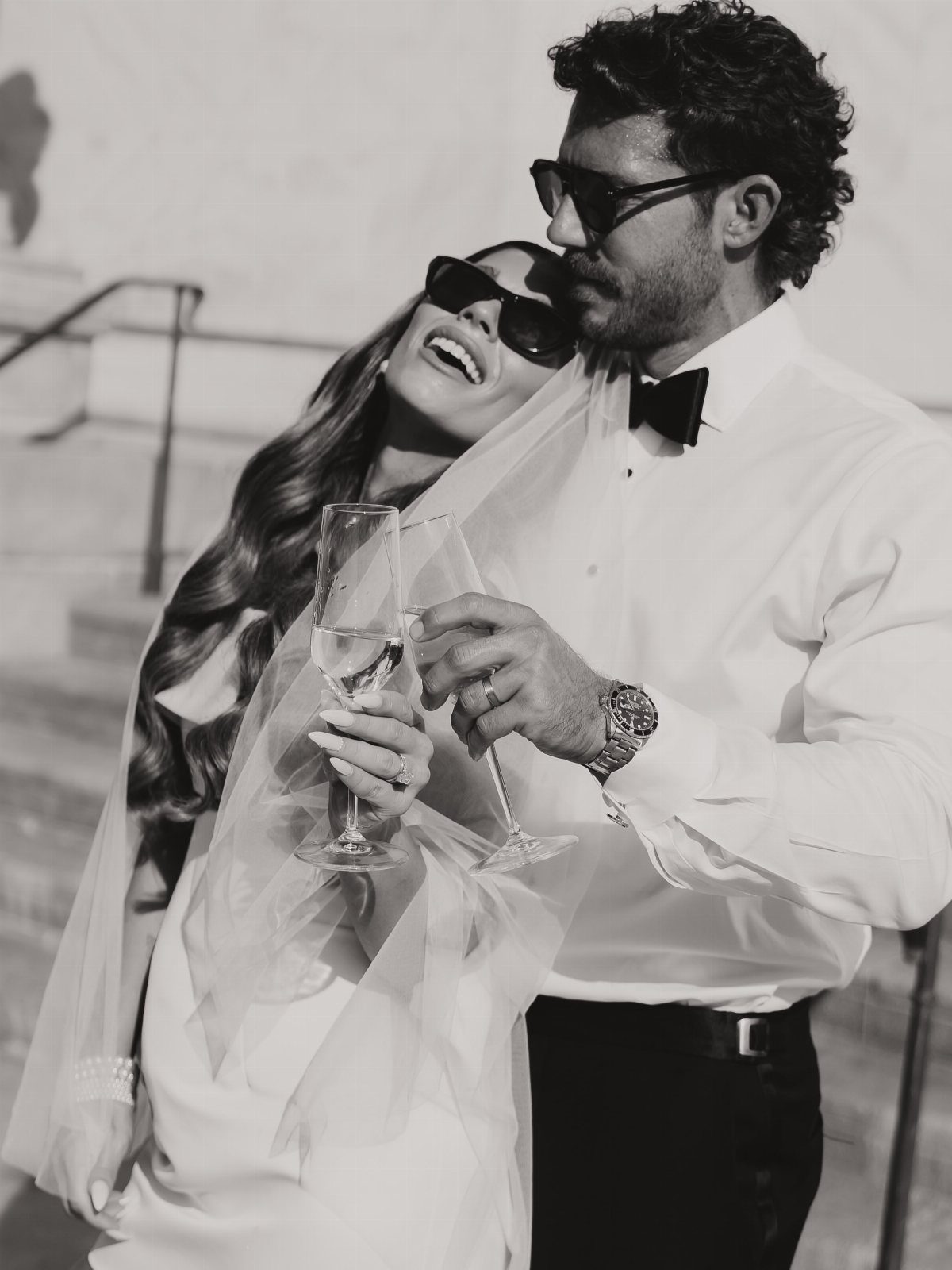 Smiling bride and groom in wedding attire wearing sunglasses and holding champagne glasses.